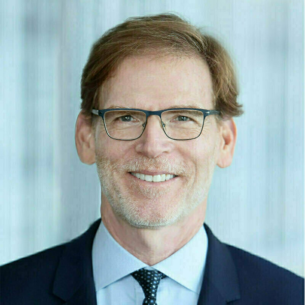 Smiling man with brown hair, goatee, and dark glasses wears a navy suit, light blue shirt, and patterned tie.